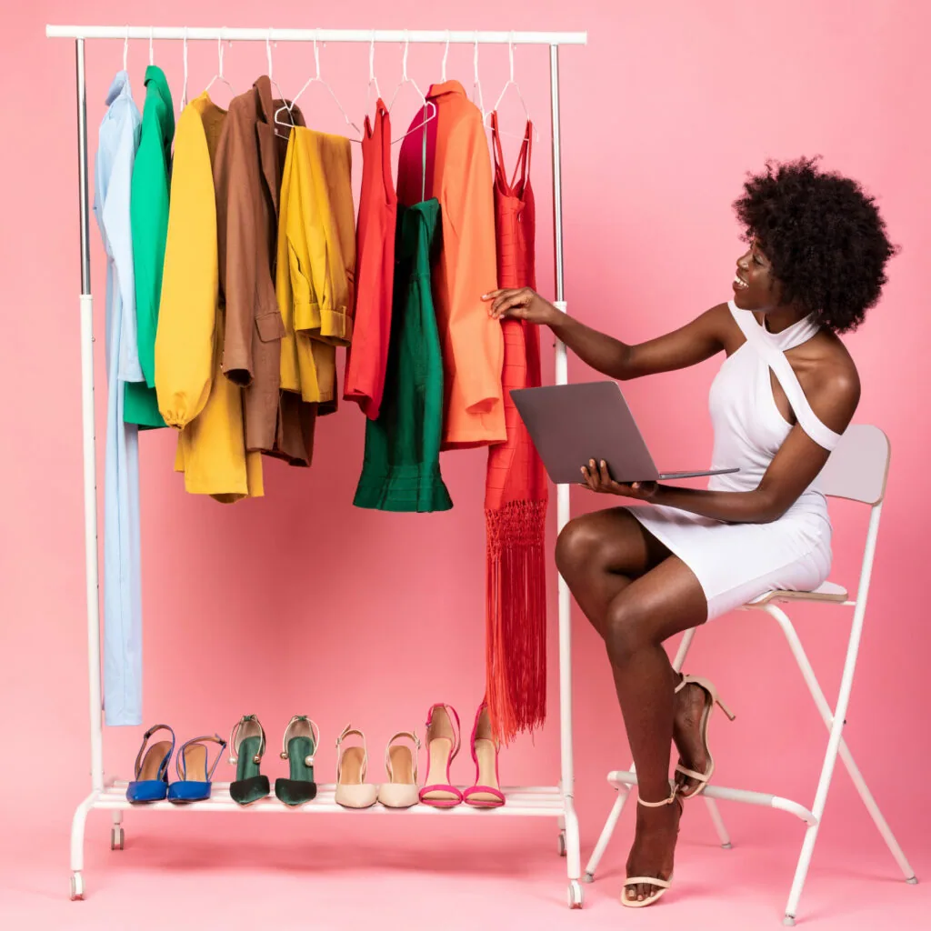 Woman in white dress sitting on folding chair holding laptop, browsing colorful clothing on white metal rack with shoes displayed below