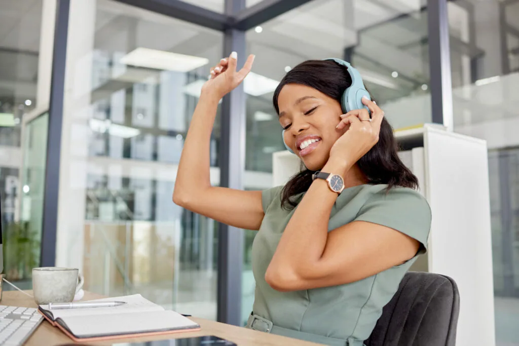 Woman wearing light blue headphones at desk, smiling with hand raised, in modern office with glass walls