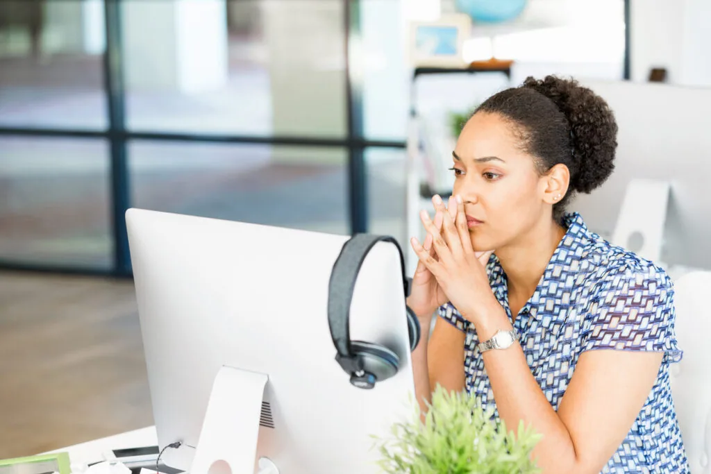 Woman in blue patterned shirt sitting at desk with computer monitor and headphones, hand near face in thoughtful pose