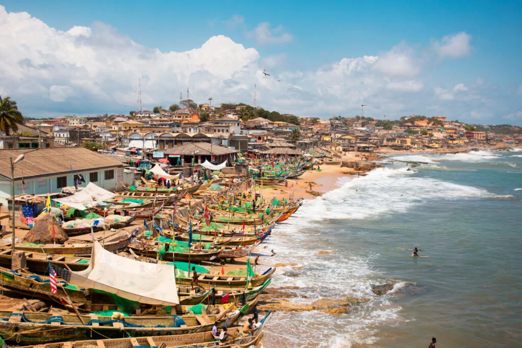 Colorful fishing boats lined up on sandy beach with hillside settlement and ocean waves in background