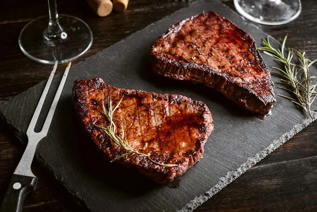 Two grilled steaks on slate plate with rosemary sprigs, fork, and wine glasses
