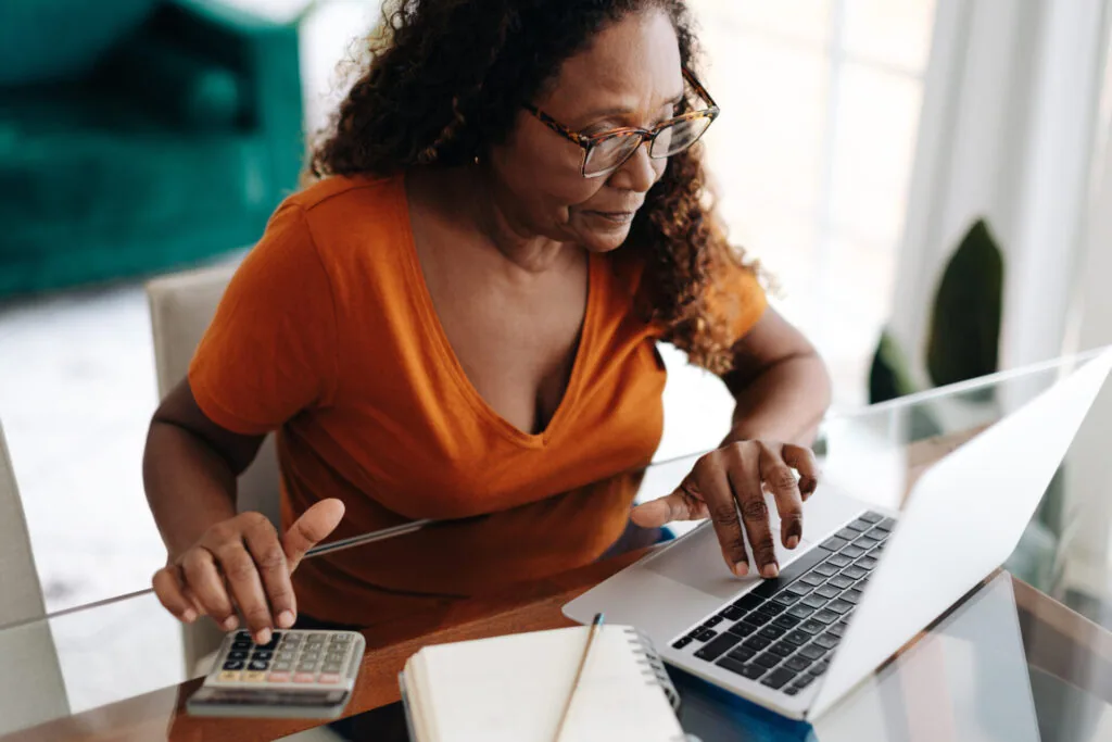 Woman in orange shirt wearing glasses working at laptop with calculator on desk