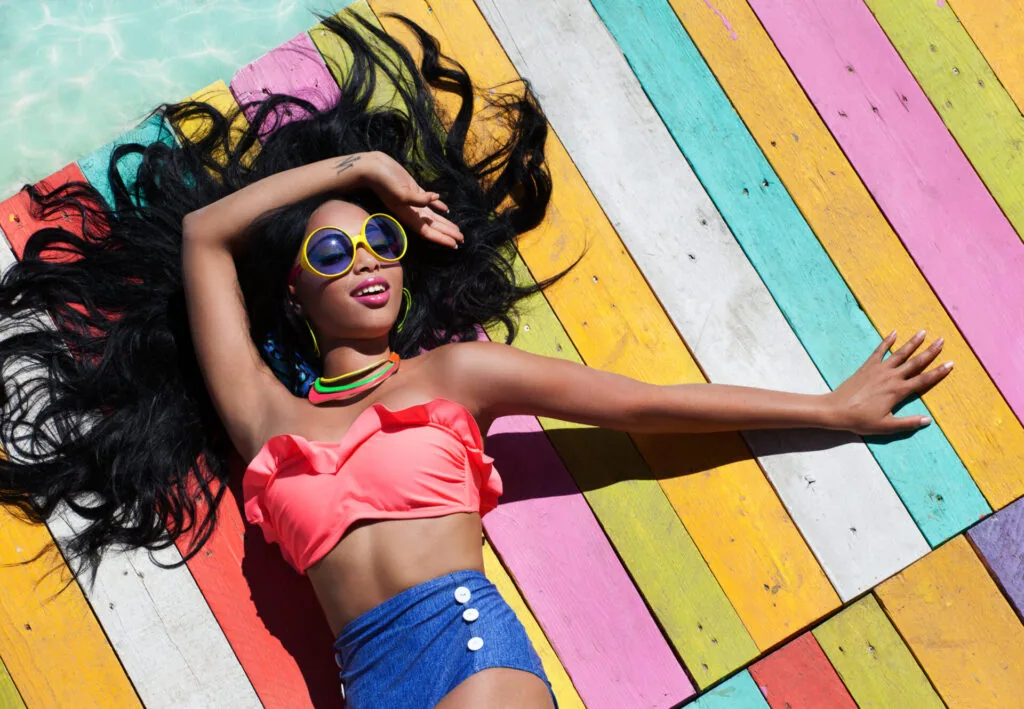 Person with long black hair wearing yellow-framed sunglasses, pink bikini top, and blue shorts lying on colorful striped wooden deck beside turquoise water