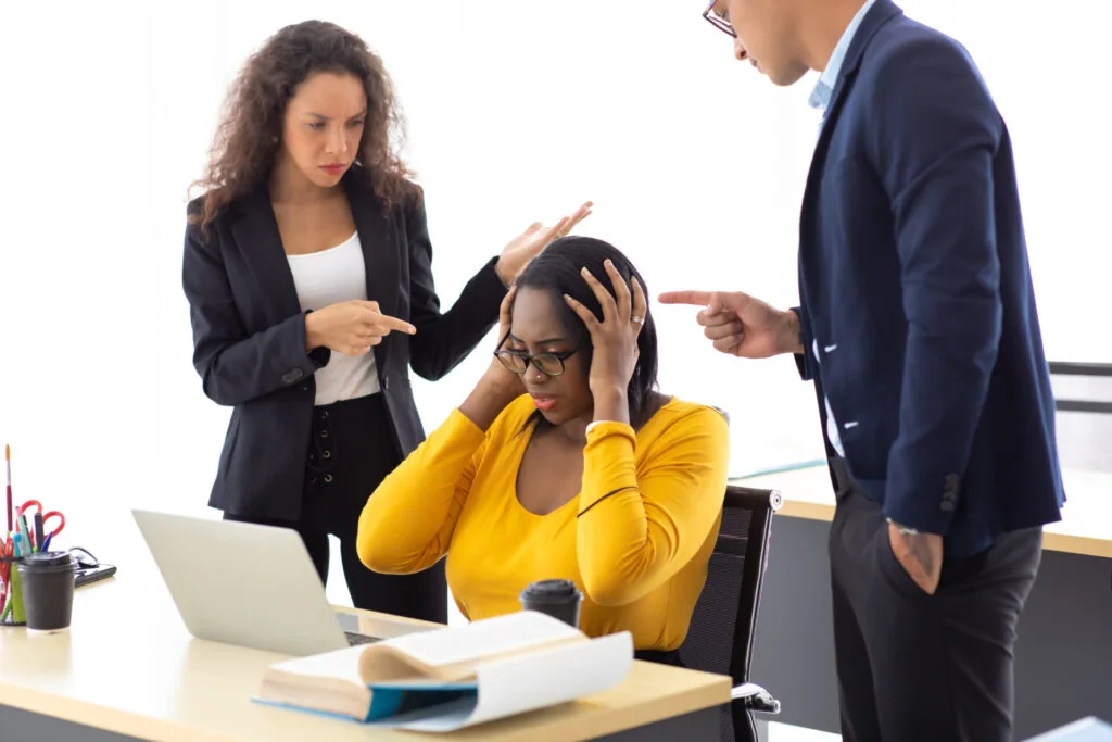 Woman in yellow shirt at desk with head in hand, two business people in navy blazers gesturing while speaking to her