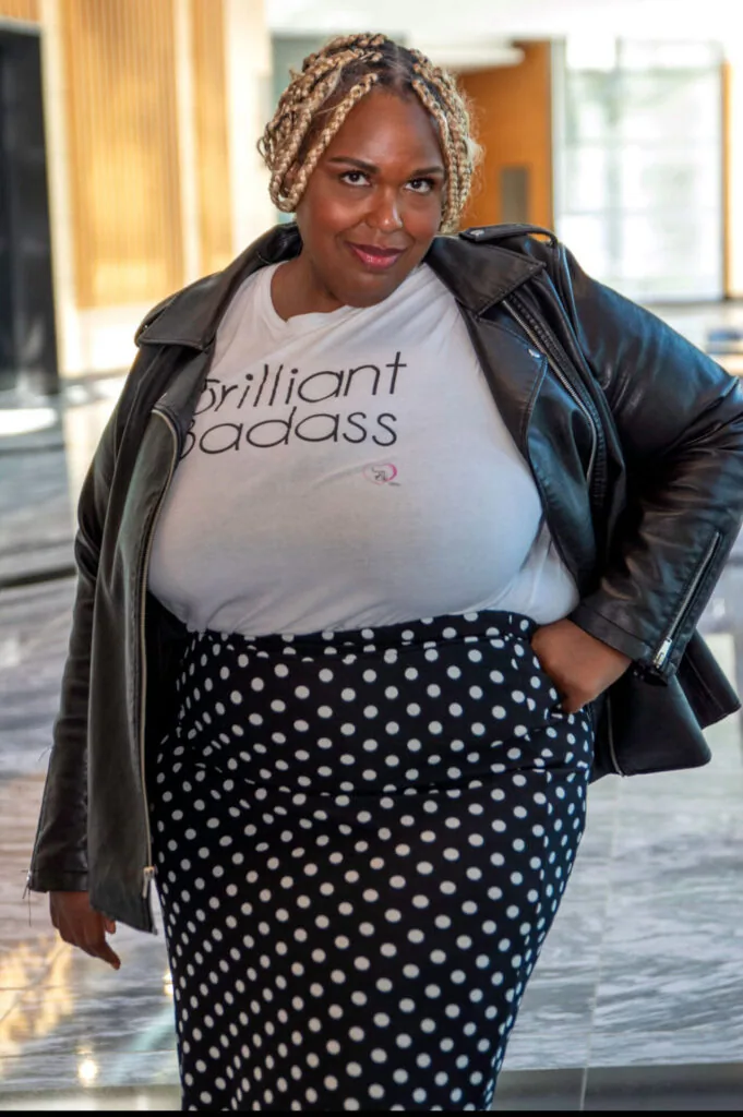 Woman wearing gray graphic t-shirt, black leather jacket, and black polka dot skirt standing indoors